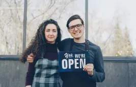 Two small business owners holding 'Yes We're Open' sign outside their storefront