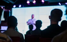 An audience watching a speaker on a bright screen in a dark room